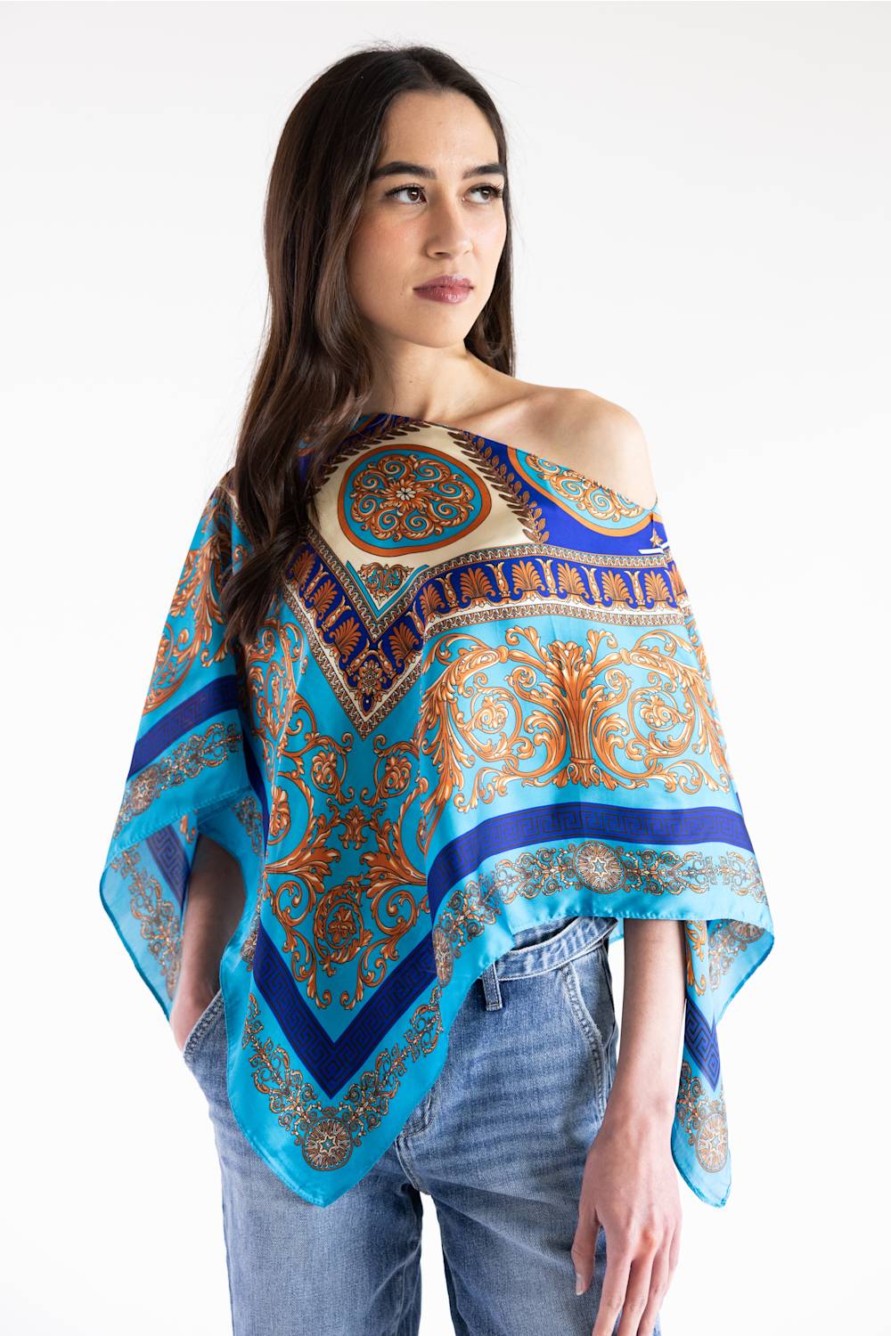 A woman modeling a vibrant turquoise and blue patterned poncho-style top with gold and brown ornate motifs, paired with blue jeans against a plain white studio background.