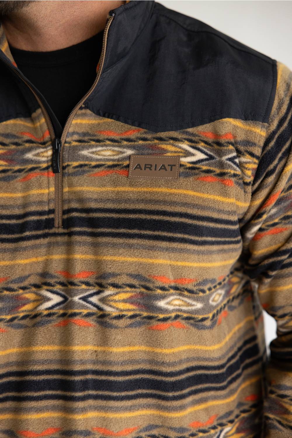 A close-up of a person wearing a quarter-zip fleece pullover with a black collar and a Southwestern geometric pattern in beige, brown, yellow, and orange. The brand name 'ARIAT' is visible on a small patch on the chest.