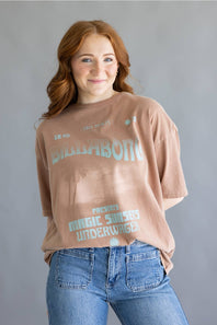 A woman with red hair wearing a faded brownish-pink Billabong t-shirt with a light blue graphic print and blue jeans, standing against a solid gray background.