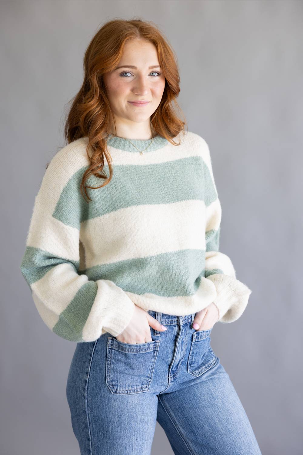 A woman with red hair wearing a fuzzy, long-sleeve knit sweater with horizontal sage green and cream stripes, paired with blue jeans, against a solid gray studio background.