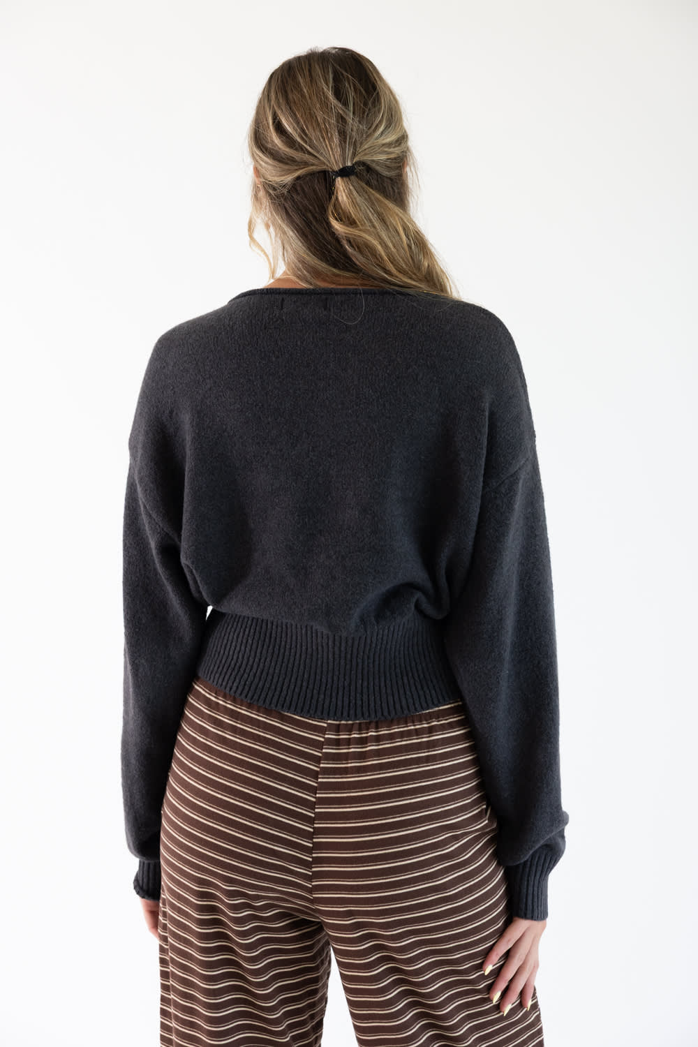 A woman viewed from behind wearing a dark charcoal gray knit sweater and brown striped pants against a plain white background.