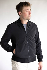 A man wearing a black zip-up bomber jacket over a gray t-shirt and off-white pants, standing against a plain light gray background.