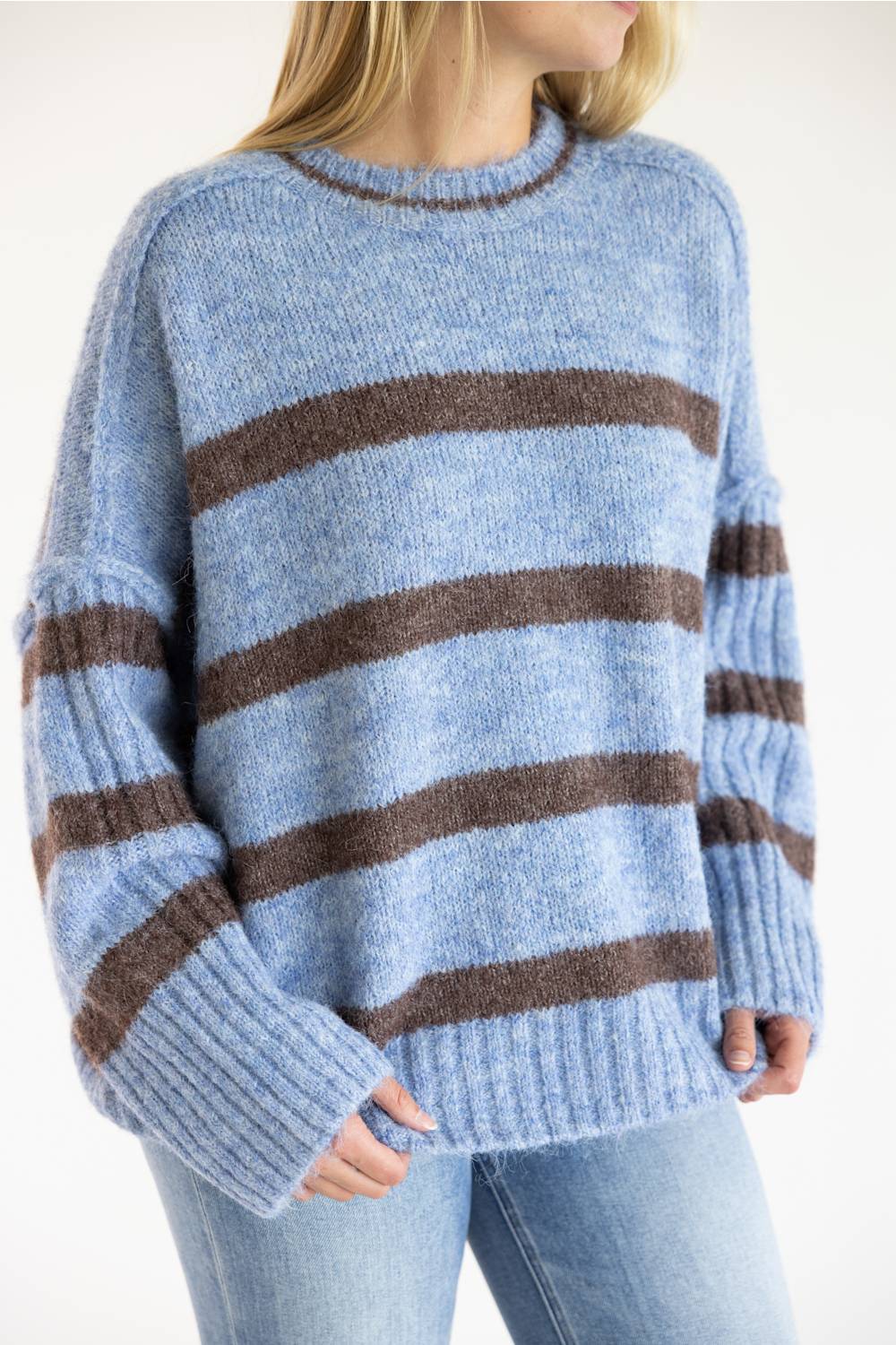 A person wearing a fuzzy knit sweater with horizontal brown stripes on a light blue base, paired with light blue jeans against a plain light background.