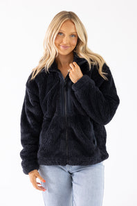 A woman wearing a black fuzzy fleece zip-up jacket and light wash jeans, standing against a plain white studio background.