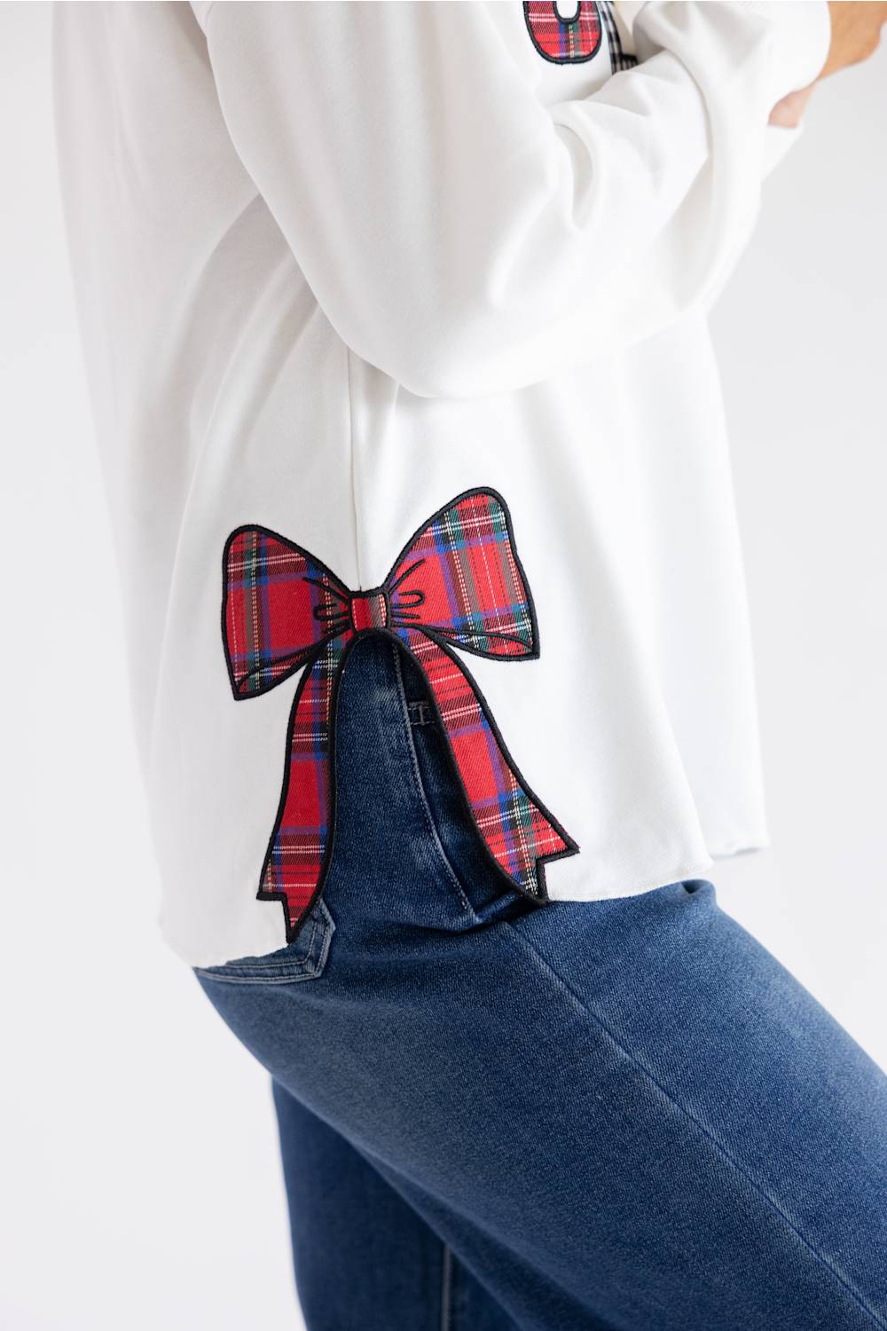 A close-up of a person wearing a white sweatshirt with a large red and blue tartan plaid bow applique on the back pocket, paired with blue jeans.