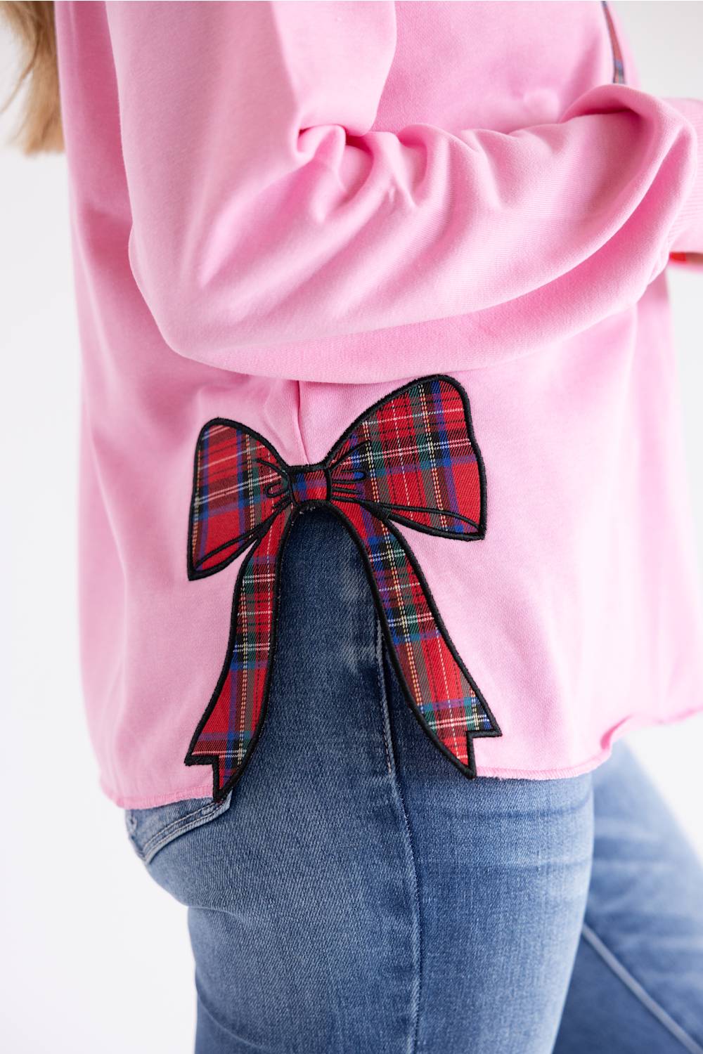 A close-up of a person wearing a pink sweatshirt with a large red and blue plaid bow applique on the back, paired with blue jeans against a white background.
