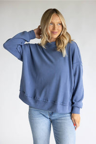 A woman wearing an oversized dusty blue sweatshirt and light-wash blue jeans, posing against a plain light gray studio background.