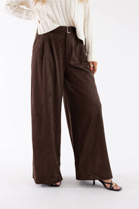 A person wearing dark brown wide-leg pleated trousers with a matching belt, styled with a cream ribbed top and black strappy heels against a plain white studio background.