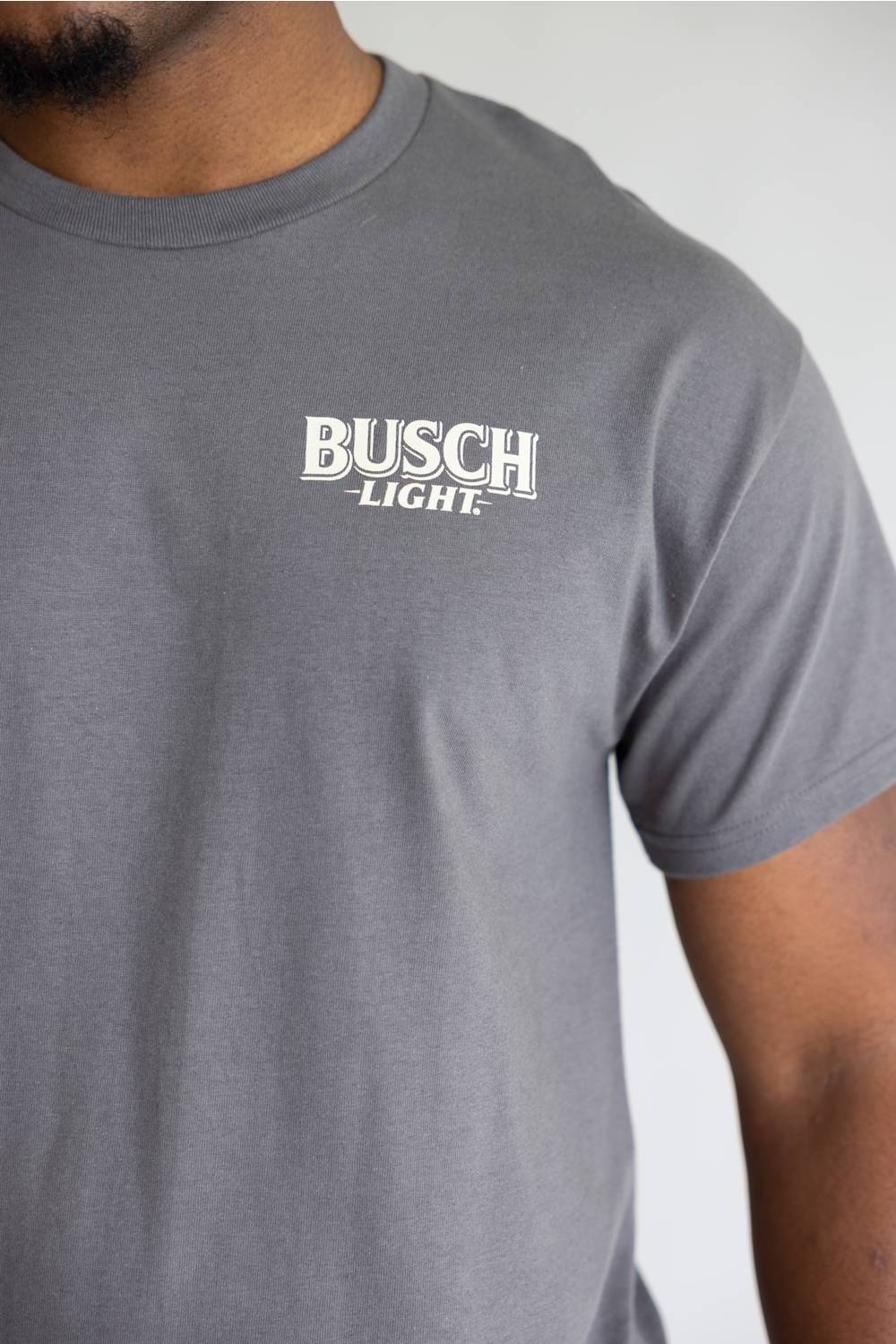 A close-up of a person wearing a heather gray crewneck t-shirt with a white 'BUSCH LIGHT.' logo printed on the left chest, against a plain light gray background.