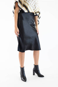 A model shown from the side wearing a black satin midi skirt with a side slit, paired with a patterned top and black ankle boots, against a plain white background.
