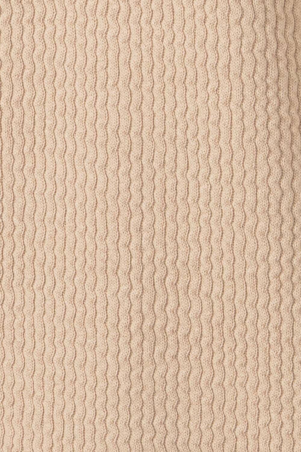 A close-up of a ribbed knit fabric in a warm beige or cream color, showing the vertical wavy texture of the yarn.