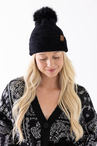 A woman looking down with a gentle smile, wearing a black plush pom-pom beanie featuring a cuffed hem with a C.C. branded leather patch, paired with a black and white patterned cardigan.