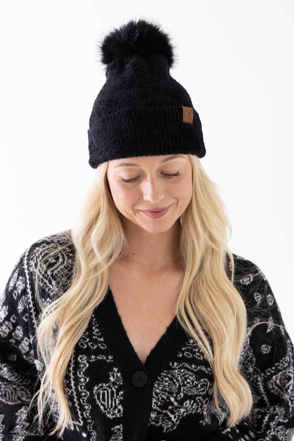 A woman looking down with a gentle smile, wearing a black plush pom-pom beanie featuring a cuffed hem with a C.C. branded leather patch, paired with a black and white patterned cardigan.