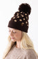 Woman wearing a brown and cream polka dot knit beanie with a fuzzy brown pom-pom and a C.C. brand patch on the cuffed hem.