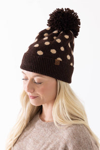 Woman wearing a brown and cream polka dot knit beanie with a fuzzy brown pom-pom and a C.C. brand patch on the cuffed hem.