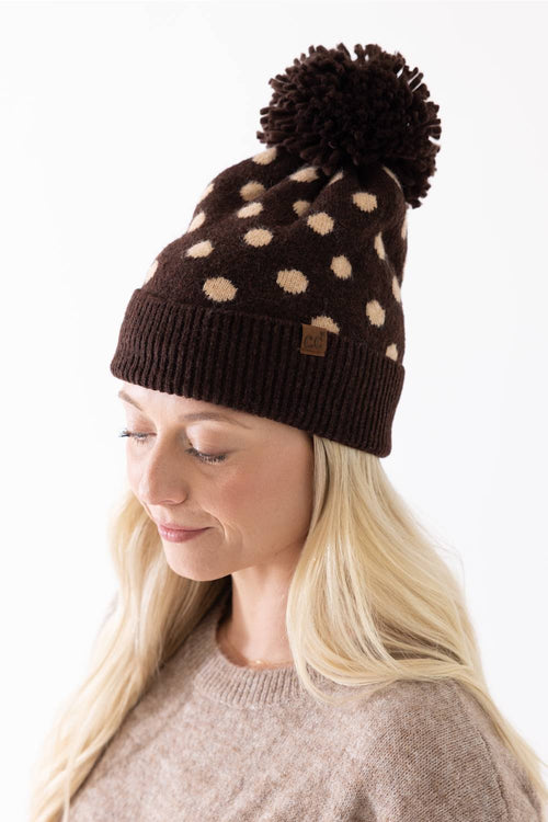 Woman wearing a brown and cream polka dot knit beanie with a fuzzy brown pom-pom and a C.C. brand patch on the cuffed hem.