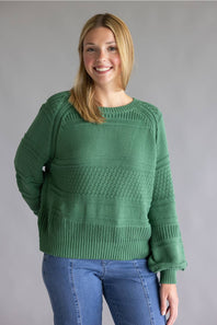 A woman wearing a green textured knit sweater and blue jeans, standing against a solid gray studio background.