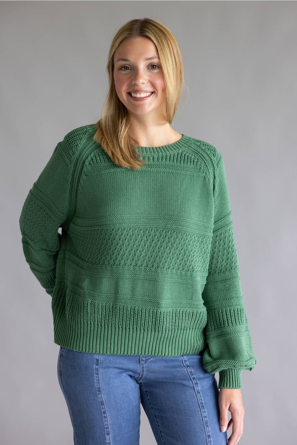 A woman wearing a green textured knit sweater and blue jeans, standing against a solid gray studio background.