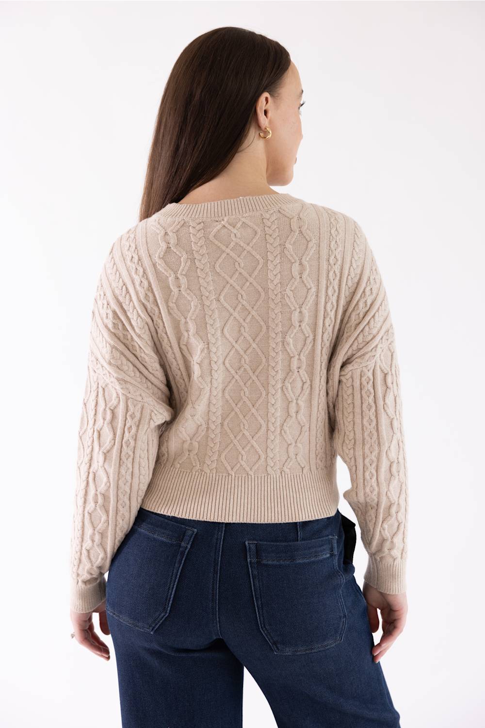 A woman shown from the back wearing a beige cable-knit sweater and dark blue jeans against a plain white studio background.