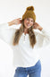 A woman wearing a mustard yellow ribbed knit beanie with a pom-pom and a white ribbed henley sweater, posing against a white studio background.