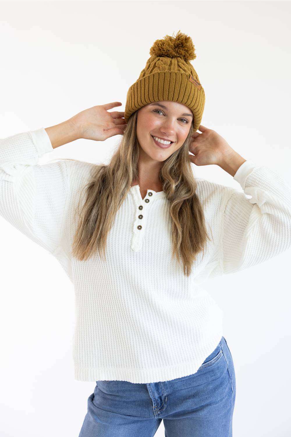 A woman wearing a mustard yellow ribbed knit beanie with a pom-pom and a white ribbed henley sweater, posing against a white studio background.