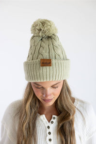 A woman wearing a sage green cable-knit beanie with a brown leather brand patch and a matching pom-pom, looking down against a plain white background.