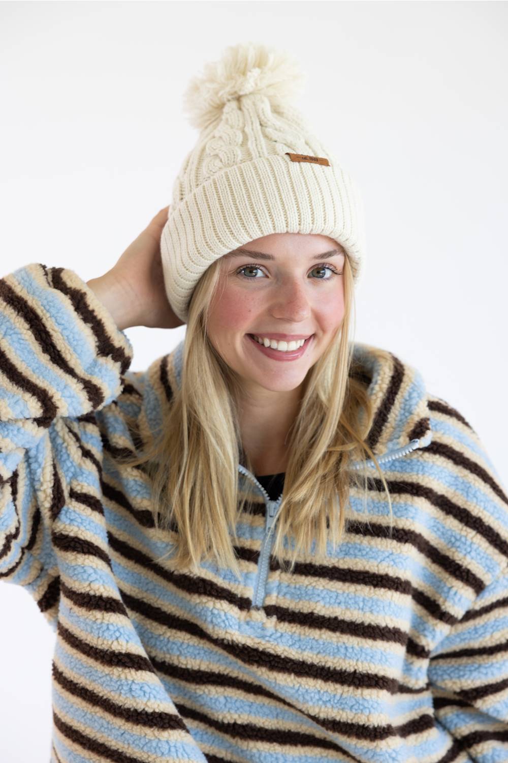 A smiling blonde woman wearing a cream cable-knit beanie with a pom-pom and a light blue, brown, and cream striped fleece pullover, photographed against a clean white background.
