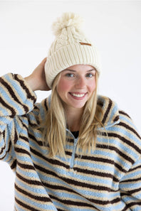 A smiling blonde woman wearing a cream cable-knit beanie with a pom-pom and a light blue, brown, and cream striped fleece pullover, photographed against a clean white background.