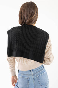 A woman shown from behind wearing a black cable-knit cropped shrug or cardigan over a cream cable-knit sweater and light blue jeans, against a plain white background.