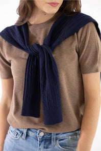A woman wearing a brown short-sleeve knit top and blue jeans, with a navy blue cable-knit sweater draped over her shoulders and tied in a knot at the front.