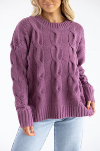 A woman wearing a chunky cable-knit crewneck sweater in a dusty mauve-purple color, paired with light-wash blue jeans, against a plain white background.