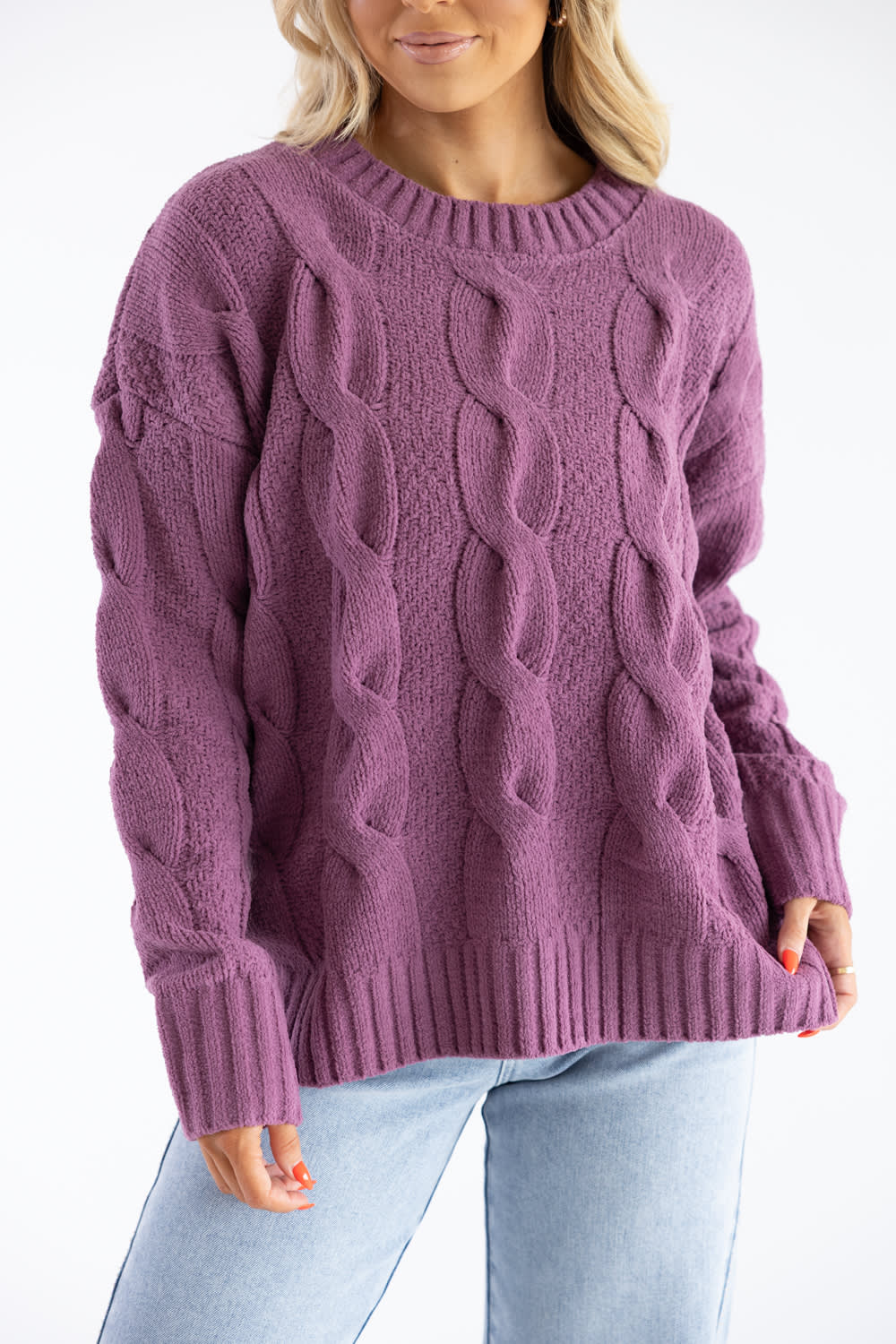 A woman wearing a chunky cable-knit crewneck sweater in a dusty mauve-purple color, paired with light-wash blue jeans, against a plain white background.
