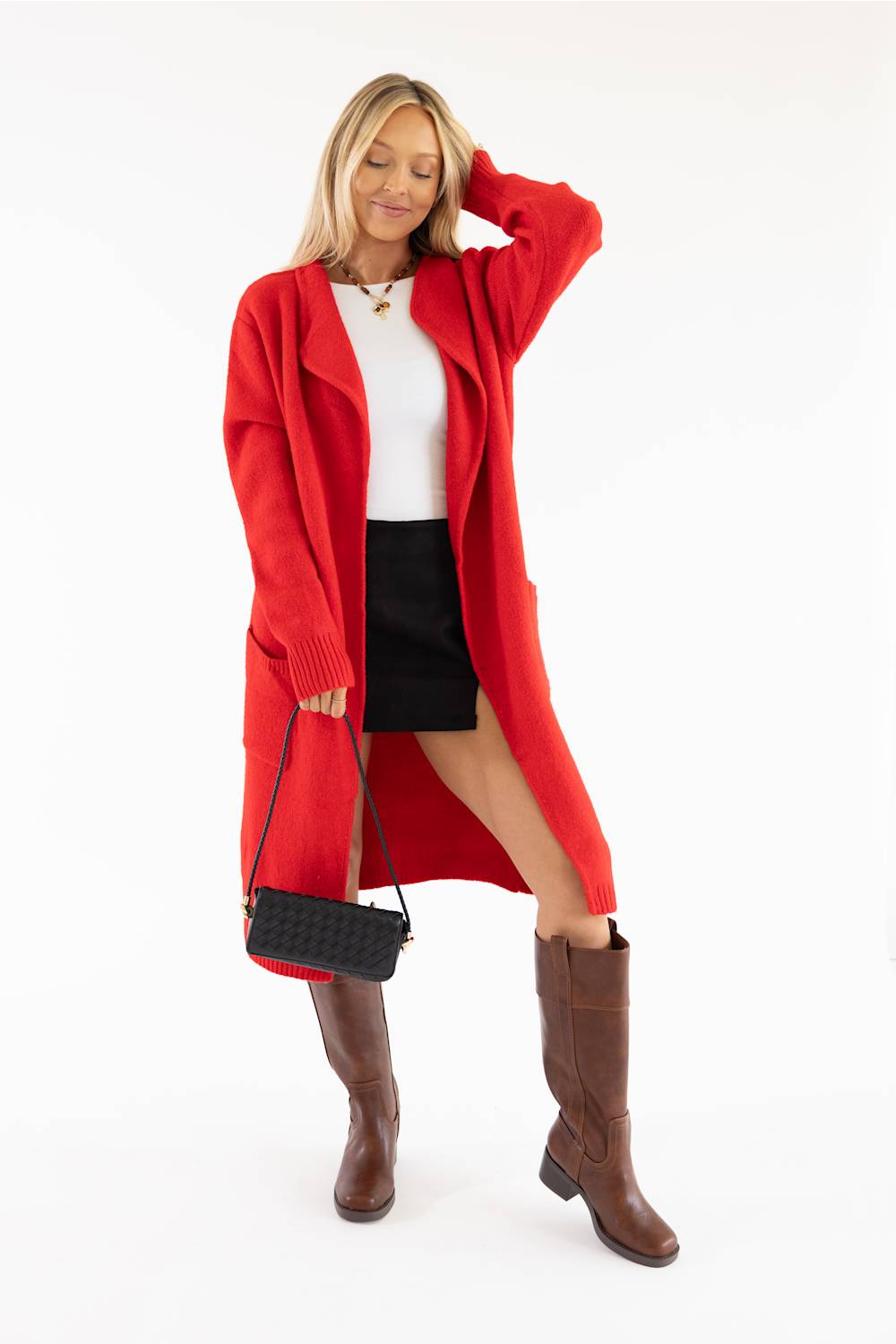 A woman modeling a bright red open-front knit cardigan over a white top and black skirt, paired with brown knee-high boots and a small black quilted shoulder bag, against a plain white studio background.