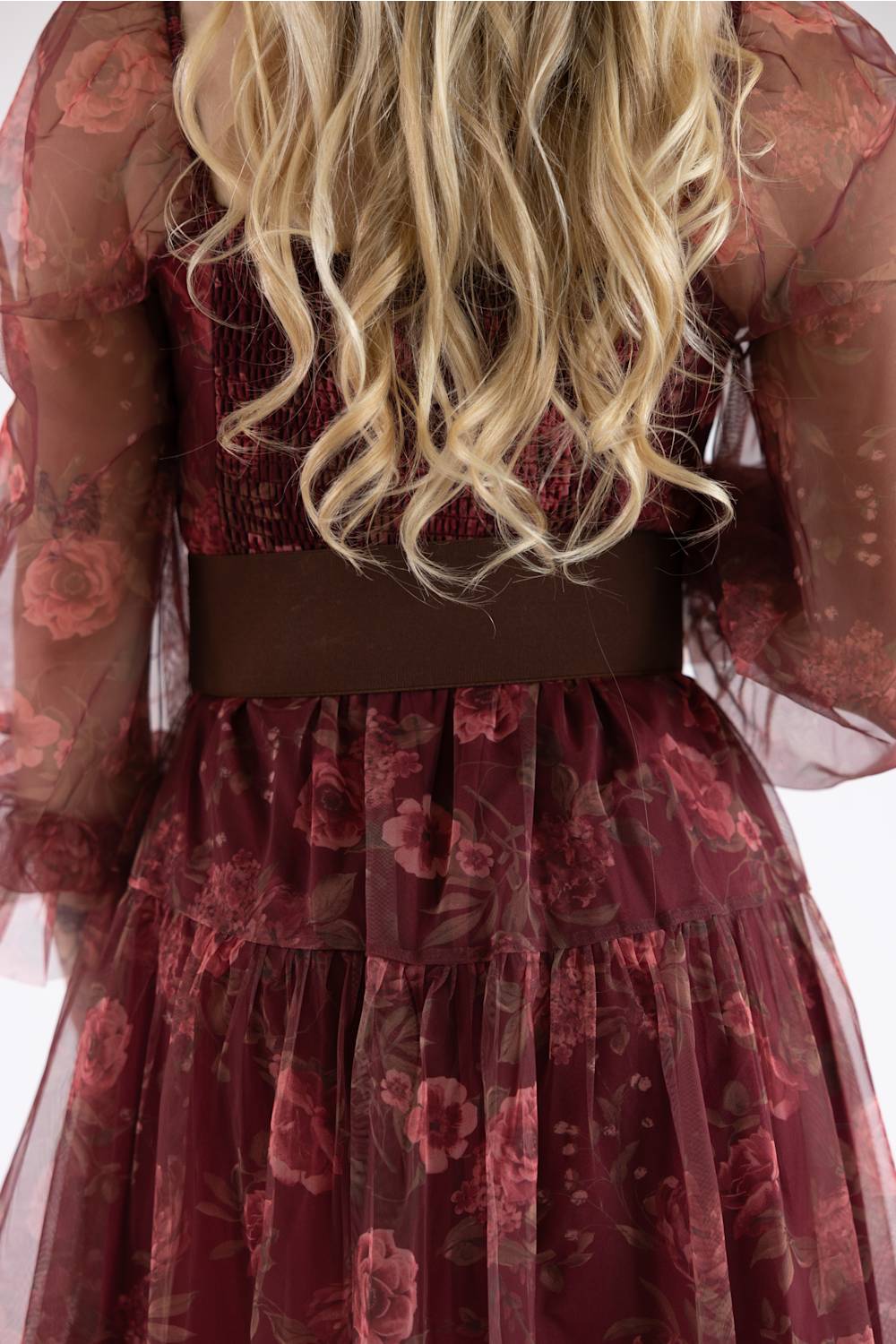 A close-up back view of a model wearing a dark burgundy tiered tulle dress with a floral pattern, sheer long sleeves, and a wide dark brown waistband.