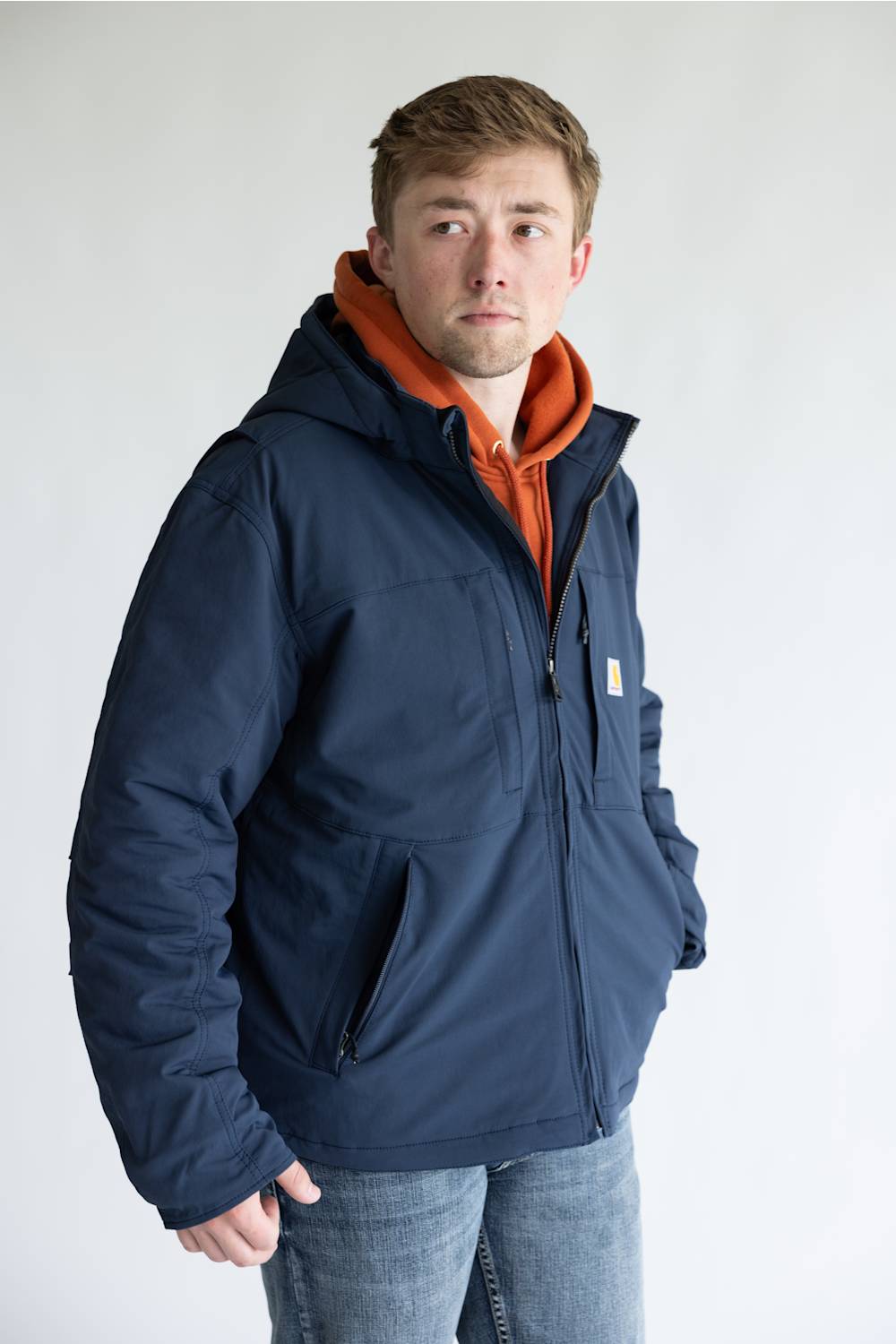 A man wearing a navy blue hooded jacket over an orange hoodie and blue jeans, standing against a plain light gray background.
