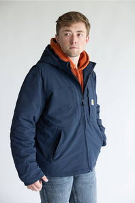 A man wearing a navy blue hooded jacket over an orange hoodie and blue jeans, standing against a plain light gray background.