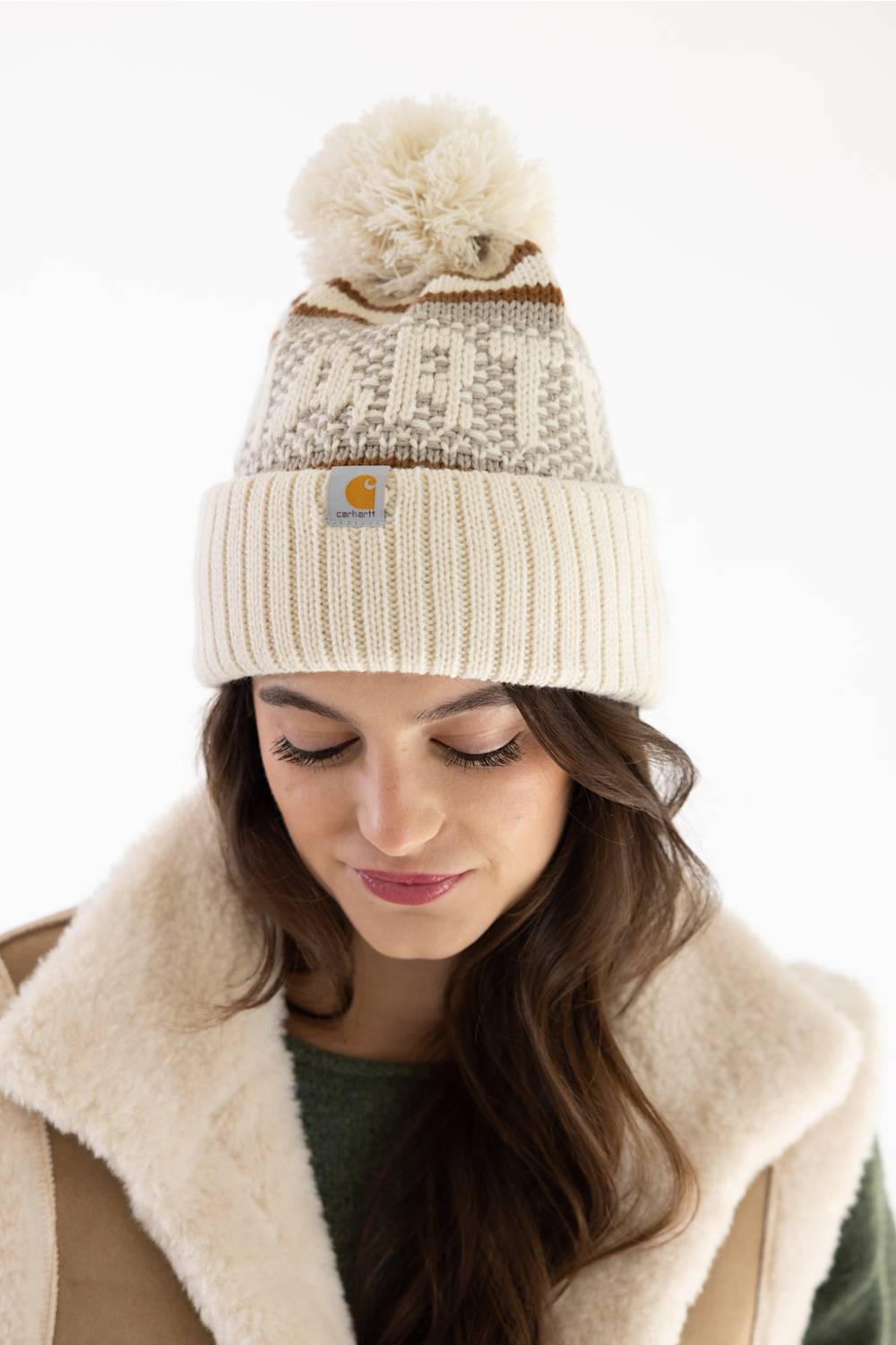 Close-up front view of a model looking down while wearing a Carhartt thick knit cuffed beanie with a pom-pom in Oat Milk and a beige sherpa vest