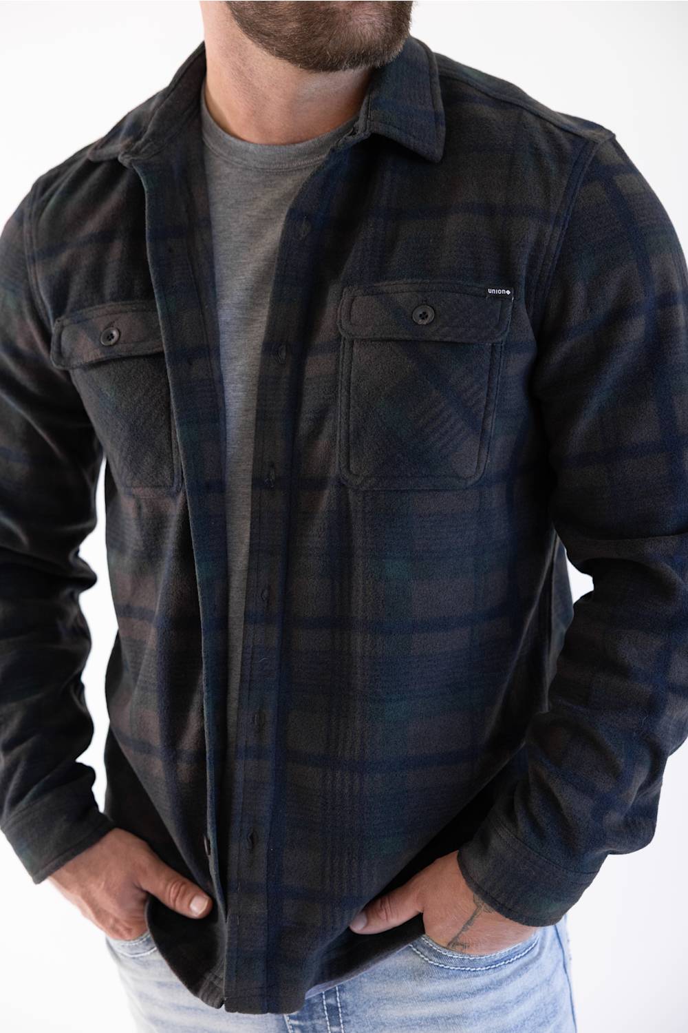 A man wearing a dark navy and black plaid button-up flannel shirt over a gray t-shirt and light blue jeans, shown from the neck down against a white background.