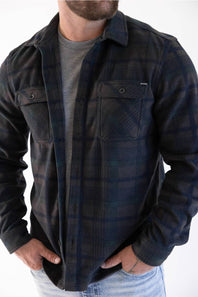 A man wearing a dark navy and black plaid button-up flannel shirt over a gray t-shirt and light blue jeans, shown from the neck down against a white background.