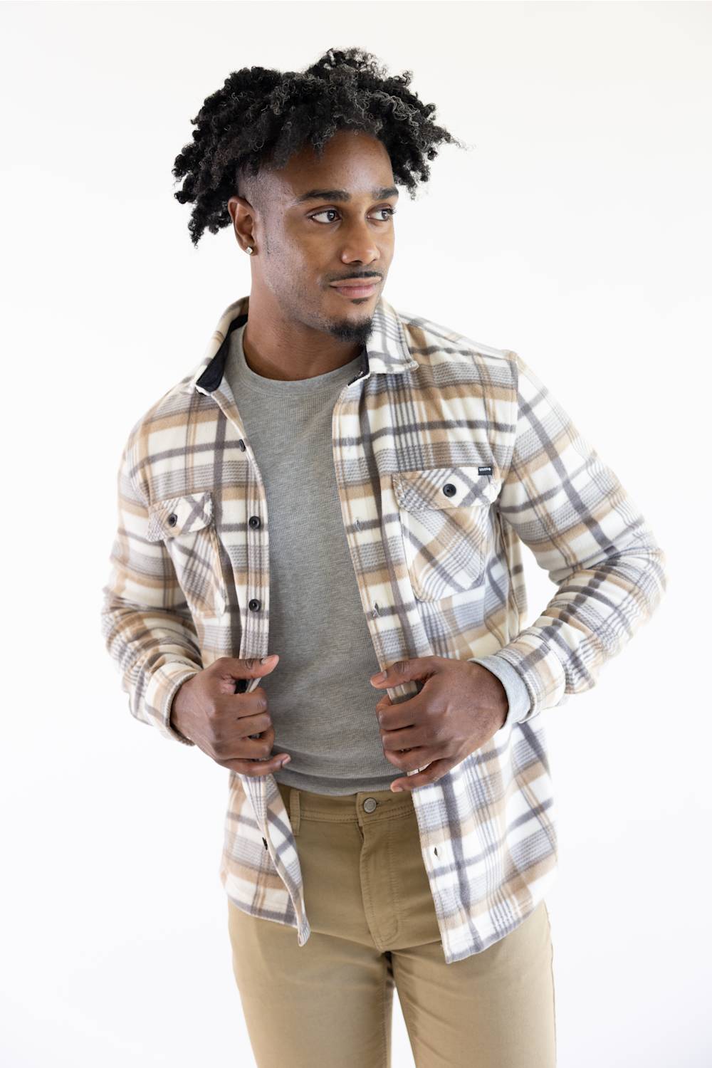 A man wearing a beige and brown plaid button-down shirt over a gray t-shirt and khaki pants, standing against a plain white studio background.