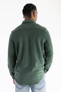 A man viewed from behind wearing a dark green long-sleeve button-up shirt and light blue jeans against a plain white background.