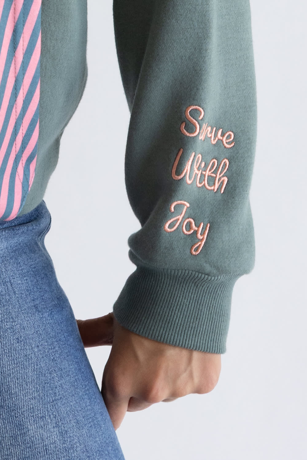 Close-up of a person wearing a sage green grey   sweatshirt with 'Save With Joy' embroidered in pink on the sleeve, paired with blue jeans and a pink striped shirt.