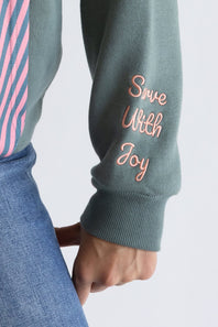 Close-up of a person wearing a sage green grey   sweatshirt with 'Save With Joy' embroidered in pink on the sleeve, paired with blue jeans and a pink striped shirt.