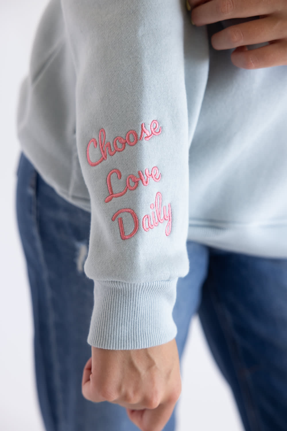 A close-up of a person wearing a light gray sweatshirt with pink embroidered text reading 'Choose Love Daily' on the left sleeve, paired with blue jeans.