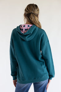 A woman viewed from the back wearing a teal green hoodie with a pink hood lining featuring black polka dots, paired with blue jeans against a plain white background.