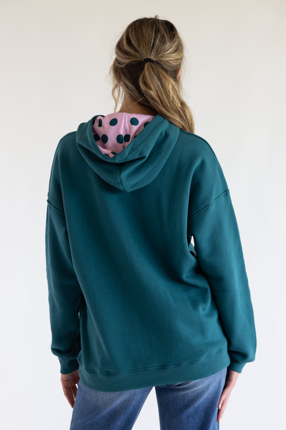 A woman viewed from the back wearing a teal green hoodie with a pink hood lining featuring black polka dots, paired with blue jeans against a plain white background.