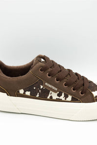 A low-top sneaker featuring brown suede and cow-print canvas panels, brown laces, and a thick white platform sole. The side of the shoe displays the brand name 'ROCKETDOG'.