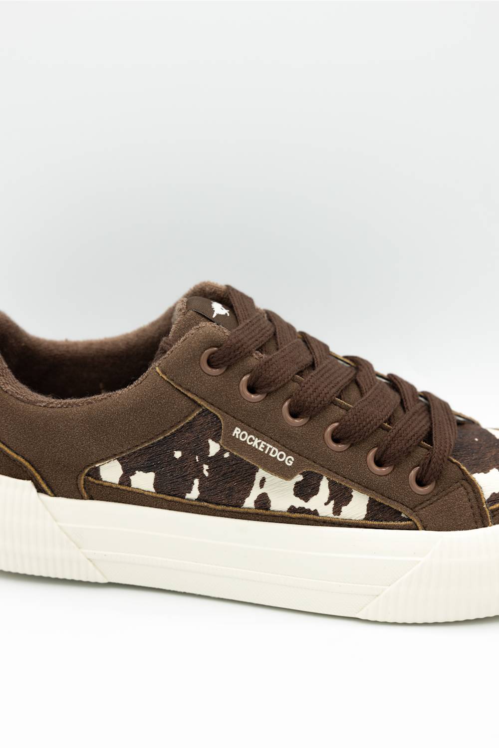 A low-top sneaker featuring brown suede and cow-print canvas panels, brown laces, and a thick white platform sole. The side of the shoe displays the brand name 'ROCKETDOG'.