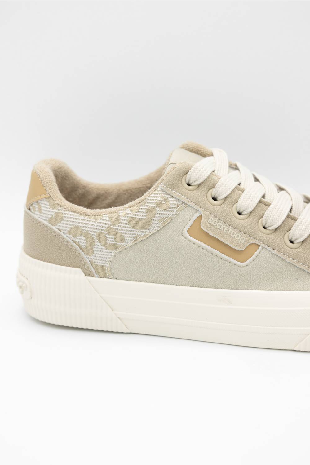 A low-top canvas sneaker in beige and off-white, featuring a textured patterned side panel, white laces, and a thick white rubber sole.