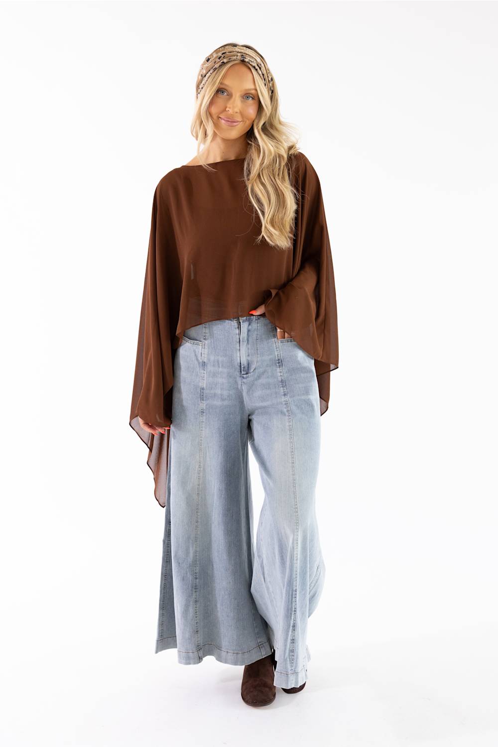 A woman modeling a brown long-sleeve top with wide bell sleeves, paired with light-wash wide-leg jeans and a patterned headband, against a plain white studio background.